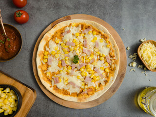 Top view closeup shot of delicious tasty juicy thin crispy cheesy corn and ham Italian pizza placed on wooden board on party table around with other ingredients, sauce, olive oil, corn and tomatoes.
