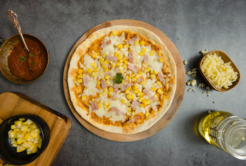 Top view closeup shot of delicious tasty juicy thin crispy cheesy corn and ham Italian pizza placed on wooden board on party table around with other ingredients, sauce, olive oil, corn and tomatoes.