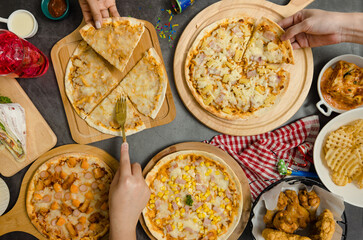 Top view shot of various kinds of delicious tasty juicy cheesy pizza placed on wooden board on party table around with other fast food while unrecognizable people picking up eating dinner together
