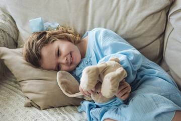Cute little girl in blue dress on the sofa at home with her favourite toy, teddy rabbit. Concept of children playing
