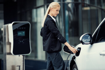 Progressive businesswoman insert charger plug from charging station to her electric vehicle with...