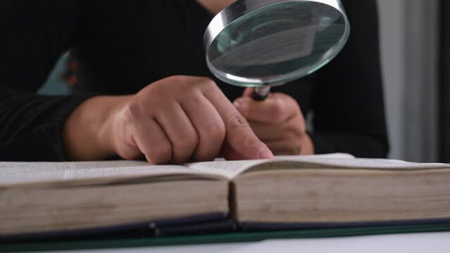 Close-up of a woman looking through a magnifying glass at a textbook. Magnifying glass in hand and open book on table. Education and research concept.