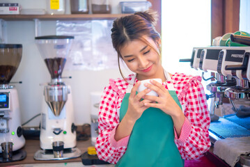 Beautiful asian barista staff wearing an apron looking at her cup of coffee meticulously brewed...