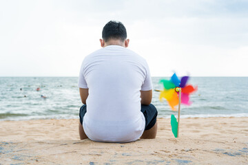 Rear view of lonely man wears a white shirt and sitting on the beach near pinwheel toy and using a...
