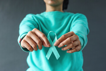 Close-up teal awareness ribbon holded by woman hands to support cancer survivor. Ovarian Cancer month, cervical cancer day.  - obrazy, fototapety, plakaty
