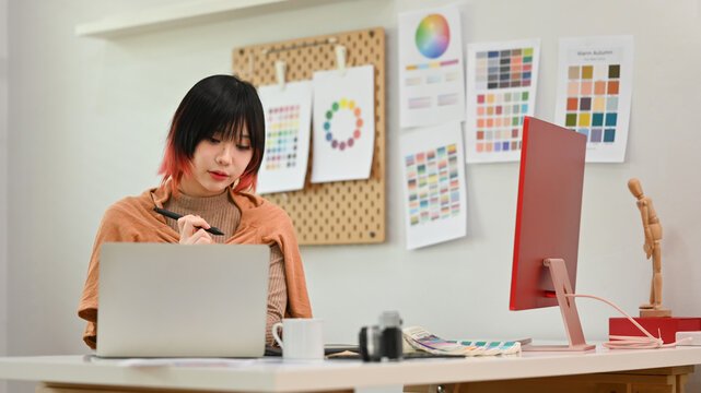 Portrait of young female designer using graphic tablet and working with color swatch samples at creative workstation