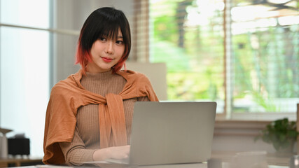 Beautiful asian woman in stylish clothes working on laptop computer at modern home office