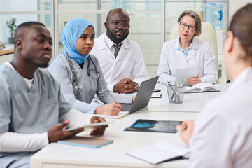 Group of doctors listening to their colleague while sitting at table during meeting in office, they...