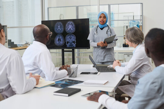 Young nurse in hijab standing near the monitor with x-ray image and presenting report to doctors during meeting in office