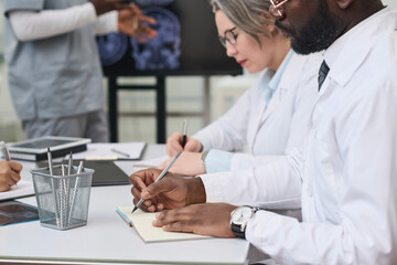 African American doctor in white coat making notes in notepad while sitting at table with his colleagues during conference