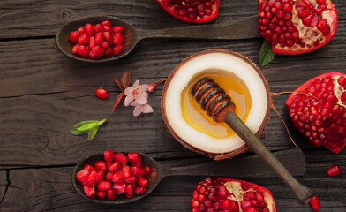 Natural liquid honey is poured into a coconut cut in half, the coconut is located on a black, old, wooden table, decorated with pieces of ripe, red, juicy, pomegranate, green mint leaves, sakura flowe