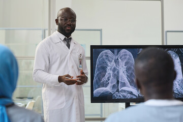 African American doctor in white coat standing near the monitor with x-ray image and discussing lungs disease with colleagues at conference