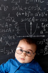 A beautiful little baby with glasses, a concept of learning and knowledge. Chalkboard with math formulas in the background.