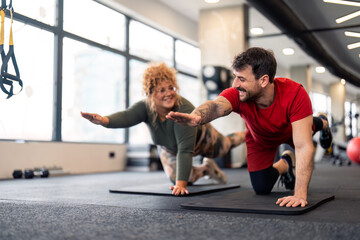 Supportive active fit man gym coach doing workout training challenging exercise for endurance, strength and stamina in gym with his fitness client positive young woman during individual fitness class.