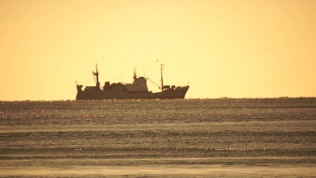 Silhouette of fishing ship in the sea on horizon. Ship sailing in the sea. Small waves on the golden surface of warm water with bokeh lights from the sun. Sea, nature and outdoor travel.