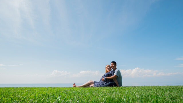 Asian man with a pregnant woman are resting in nature in a picturesque place, sit on the green grass
