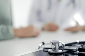 Doctor and patient are sitting and discussing something at the desk in the clinic office. The focus is on the stethoscope lying on the table, close up. Perfect medical service and medicine concept