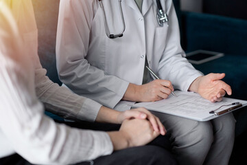 Doctor and patient sitting at sofa in clinic office. The focus is on female physician's hands filling up the medication history record form, close up. Medicine concept