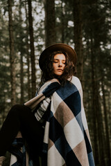 Stylish hipster girl in a hat wrapped in a checkered plaid posing in the base of a pine forest.Travel concept