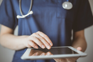 Doctor woman using tablet computer while standing near panorama window in clinic, close up. Physician or surgeon at work. Medicine concept