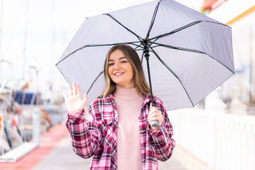 Young pretty Romanian woman holding an umbrella at outdoors saluting with hand with happy expression
