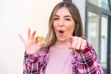 Young pretty Romanian woman holding a Bitcoin at outdoors surprised and pointing front