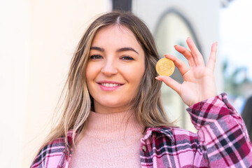 Young pretty Romanian woman holding a Bitcoin at outdoors smiling a lot