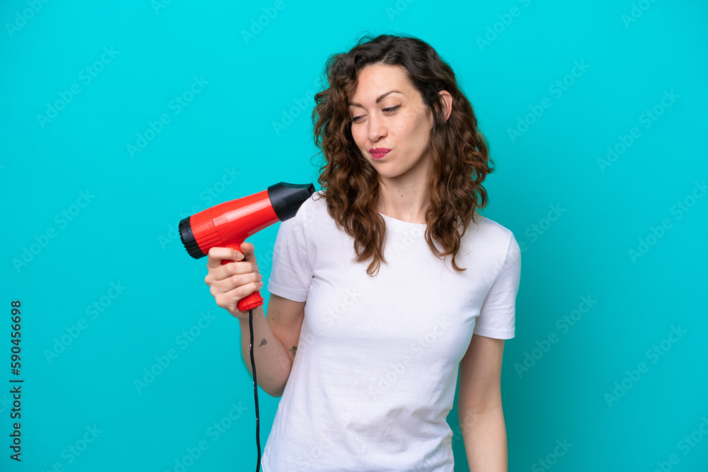 Wall mural Young caucasian woman holding a hairdryer isolated on blue background with sad expression