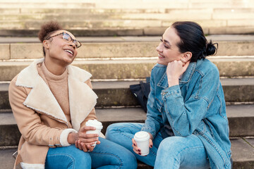 two student women in a denim jacket is talking to each other, on phone, laughing, drinking coffee  on stairs in city.