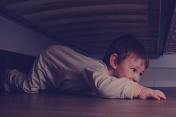 Toddler baby crawls and hides under the bed. Child climbed under the sofa and sits on the floor....