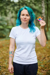 Young woman with turquoise dyed hair in white T shirt holds handmade colorful eye amulet on green...