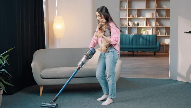 young atractive girl with a battery vacuum cleaner in the room holding little dog in her arms, cleaning. Fun cleaning in the apartment