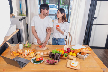 Young Asian couple cooking with fruits and vegetables and using laptop in the kitchen To cook food together within the family happily, family concept.