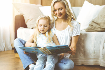 Happy family. Blonde young mother reading a book to her cute daughter while sitting at wooden floor in sunny room. Motherhood concept