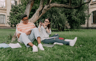 two friendly students man and woman of multiethnic students working on homework together sitting on the lawn near the tree on campus