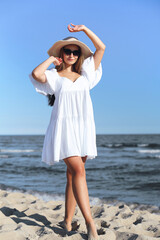 Happy blonde woman is on the ocean beach in a white dress and sunglasses, raising hands