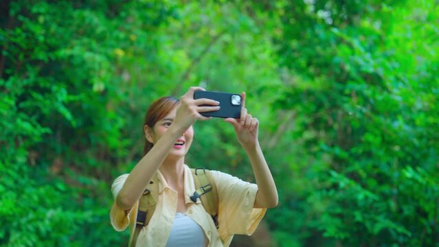 Young asian traveller using phone to take photo while walking at forest. She enjoying with backpack or camping trip alone.