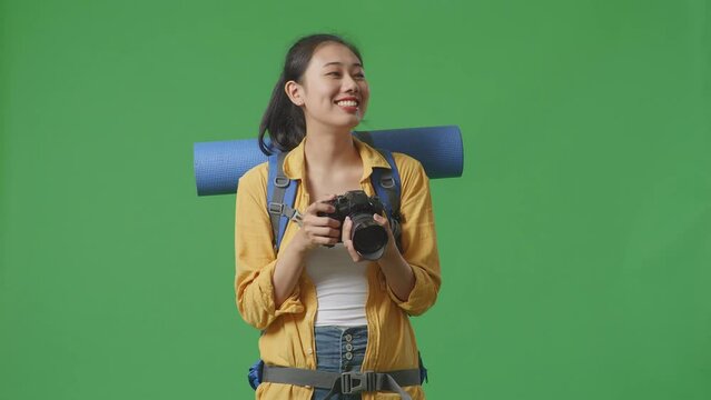 Asian Female Hiker With Mountaineering Backpack Smiling And Holding A Camera In Her Hands Then Looking Around While Standing On Green Screen Background In The Studio
