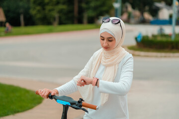 A young stylish Muslim woman in hijab looking at smart watch monitor while drives an electric scooter in the city street.