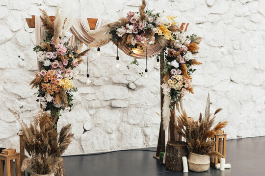 Wedding wooden arch in rustic style decorated with grass hay field coluor and flowers. Near arch is wooden baskets with flower bouquets. Wooden arch and flower decoration. Wedding ceremony