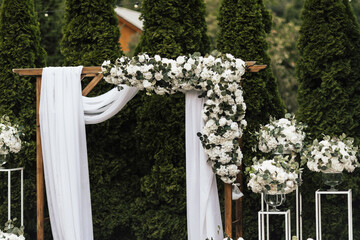 A beautiful wooden wedding arch decorated with white flowers and greenery, stands in the garden against the background of tui trees.