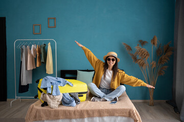 Laughing young woman sitting on bed with suitcase at home, holding hands apart in flight ready for...