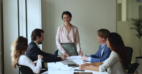 Happy mature group leader talking to team, laughing, having fun, standing at meeting table, brainstorming on project with colleagues. Corporate coach starting training with joke