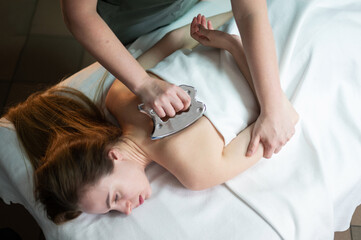 Hands of the masseur are close-up using a metal massage tool to massage the body of a woman lying on a couch. Health concept, body care, skin care, wellness