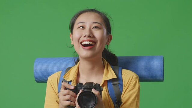 Close Up Of Asian Female Hiker With Mountaineering Backpack Smiling And Holding A Camera In Her Hands Then Looking Around While Standing On Green Screen Background In The Studio
