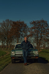 Man stands in front of a vintage american muscle car in sunny countryside.