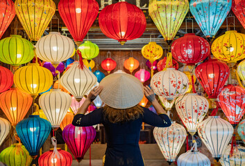 Fototapeta na wymiar Back view anonymous woman in dress touching Asian conical hat on head while standing near colorful lanterns hanging on ceiling