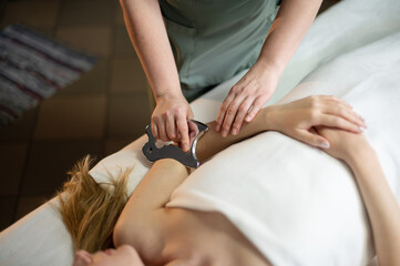 Hands of the masseur are close-up using a metal massage tool to massage the body of a woman lying...