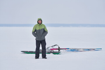 A middle-aged man, a snowsurfer, stands on the ice of a frozen lake and waits for a woman. Preparing to ride a sailboard in the snow on a cloudy winter day.
