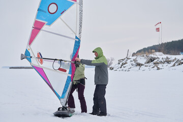 A man, a snowsurfer, helps a woman, his wife, start riding a sailboard. A middle-aged man and woman go snowsurfing on a cloudy winter day.